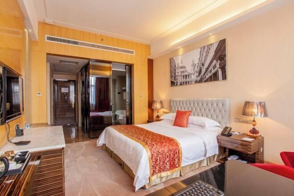 A hotel room with a bed, bedside tables, a desk, a chair, a TV, and a door leading to another room.