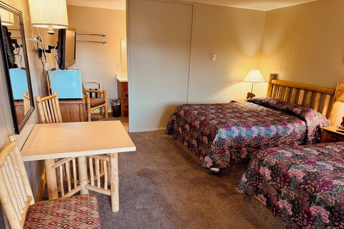 A room with a wooden table and chairs, a bed with floral bedding, a mirror, and a lamp.