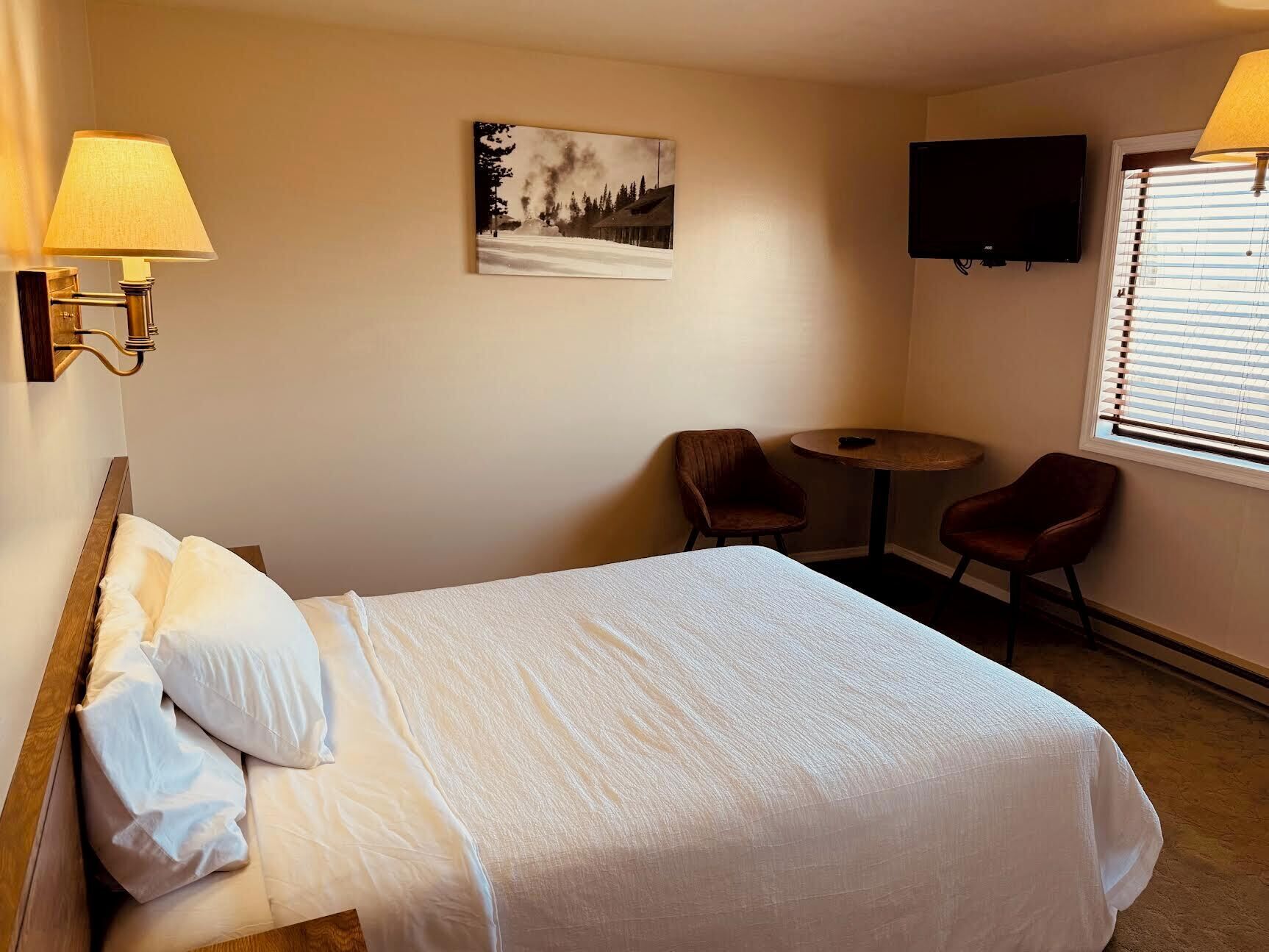 A hotel room with a bed, two chairs, a small table, a painting, a TV, and a window with blinds.