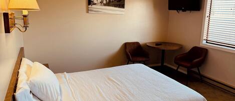 A hotel room with a bed, two chairs, a small table, a painting, a TV, and a window with blinds.