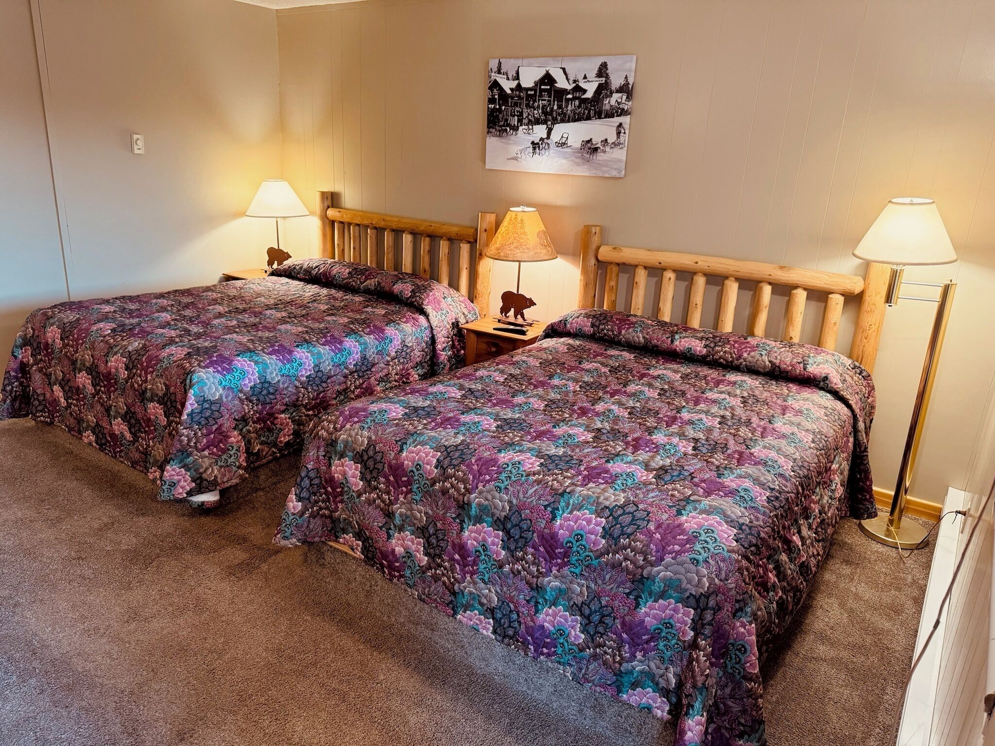 A room with two beds, each with a floral-patterned bedspread, a wooden headboard, and two bedside lamps.