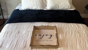 A neatly made bed with white bedding, a dark blue throw, and a wooden tray with the text "enjoy the little things" on it.
