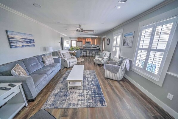 A spacious living room with a grey sofa, a white coffee table, and a patterned rug. There are two armchairs and a dining area with a kitchen in the background.