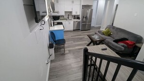A modern kitchen with white cabinets, a built-in oven, and a refrigerator. There is a dining area with a table and chairs, and a gray sofa with cushions.
