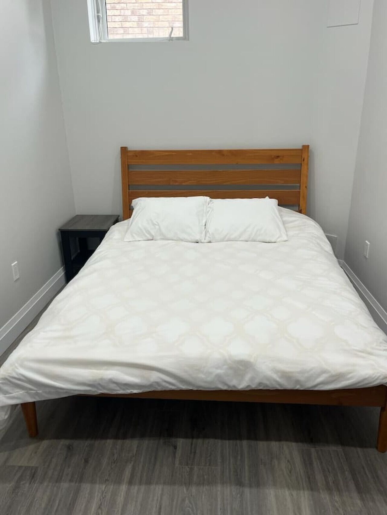 A single bed with a wooden headboard, a small bedside table, and a window with blinds.