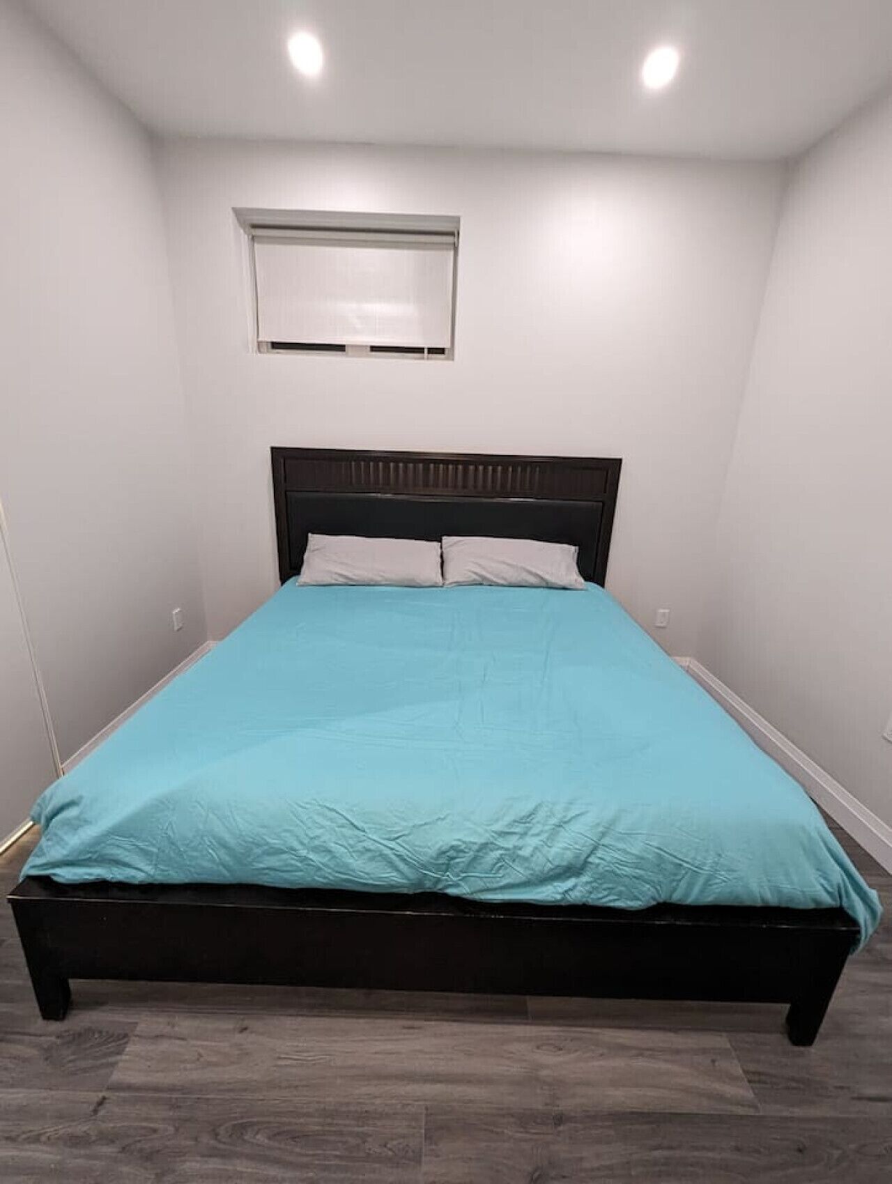 A single bed with a dark headboard, a light blue bedspread, and a small window above the bed.