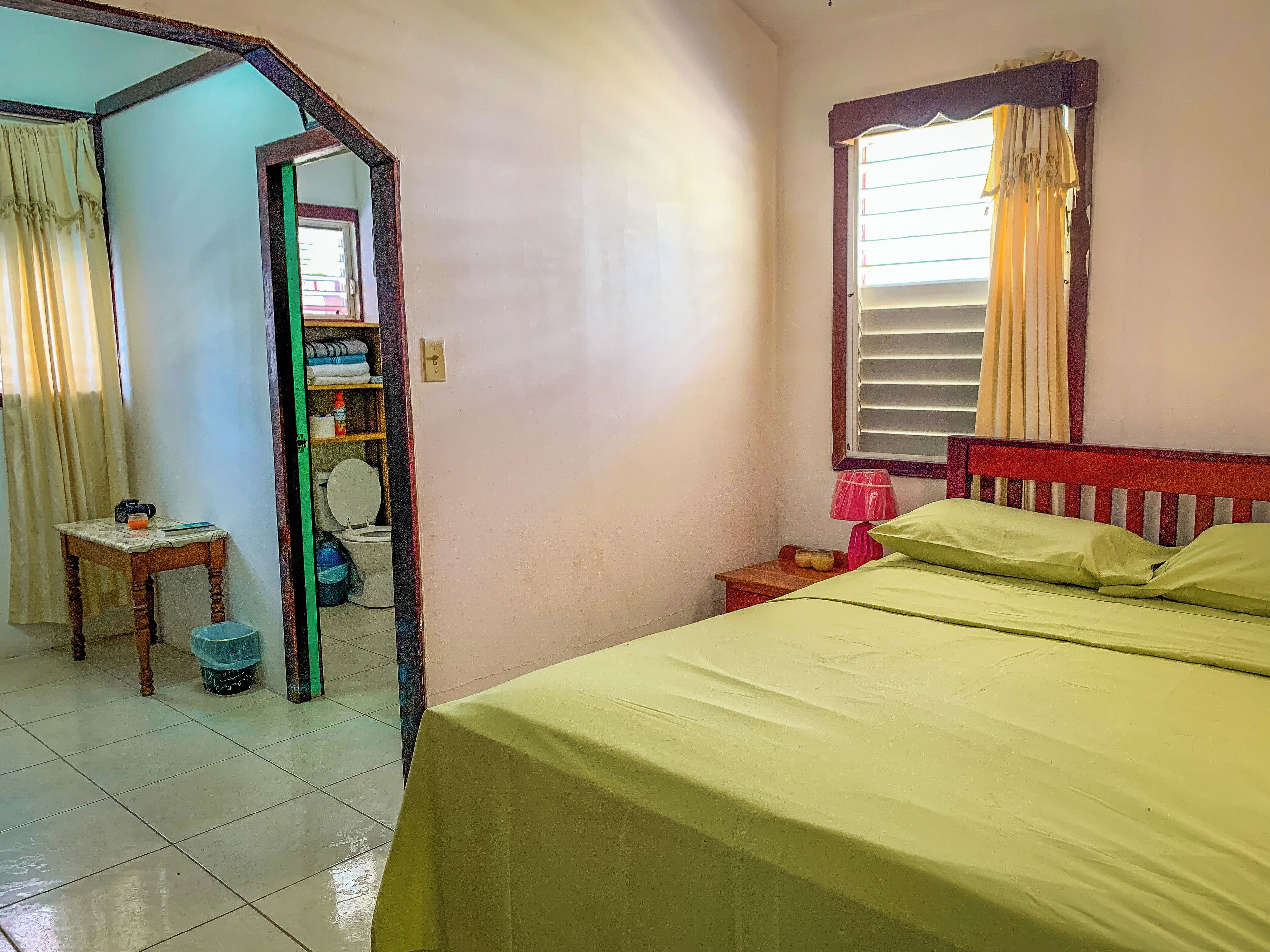 A bedroom with a bed, a nightstand, a window with curtains, a mirror, and a bathroom visible through an open door.