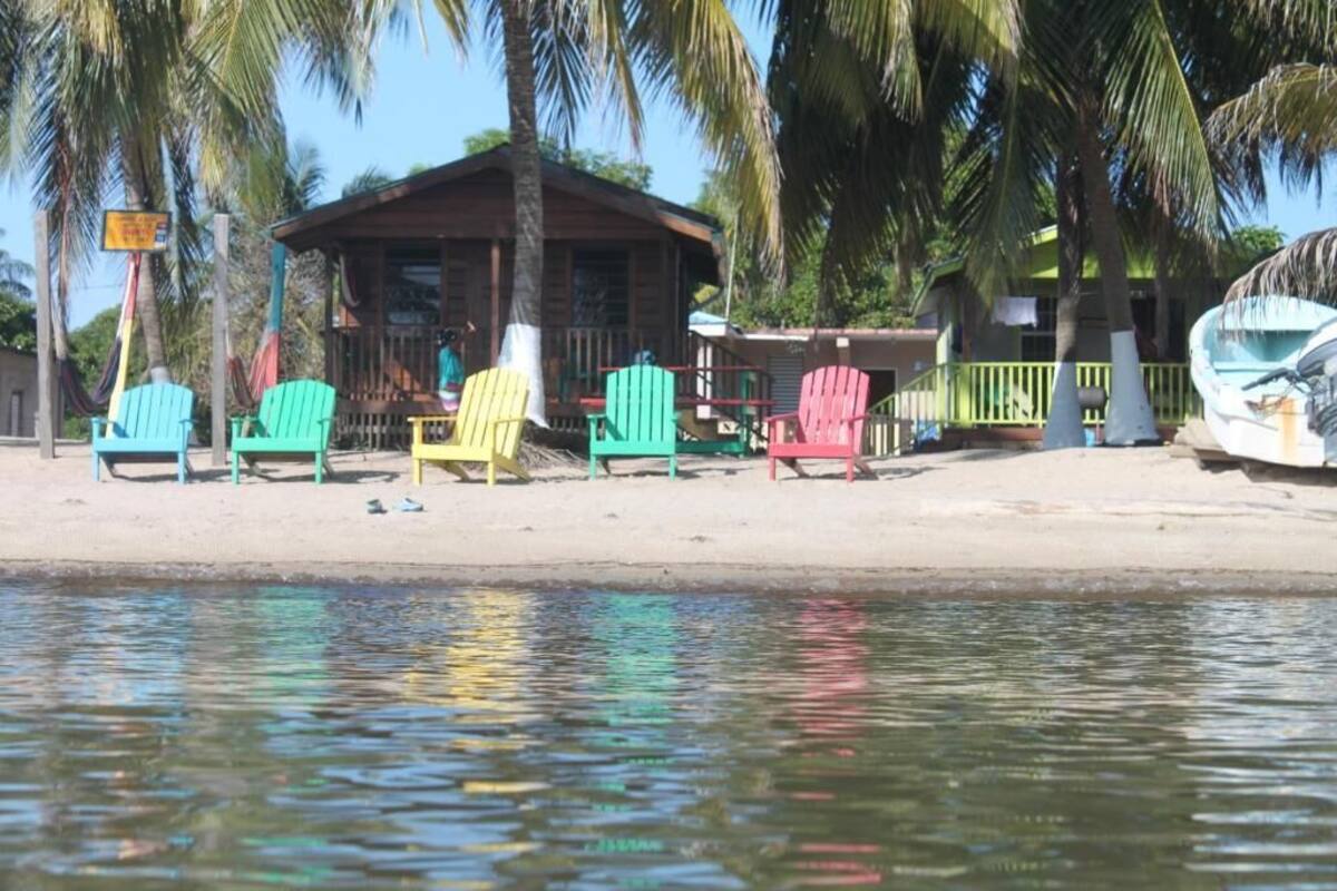 Una cabaña frente a la playa con sillas Adirondack de colores, un pequeño porche y un barco fondeado cerca.