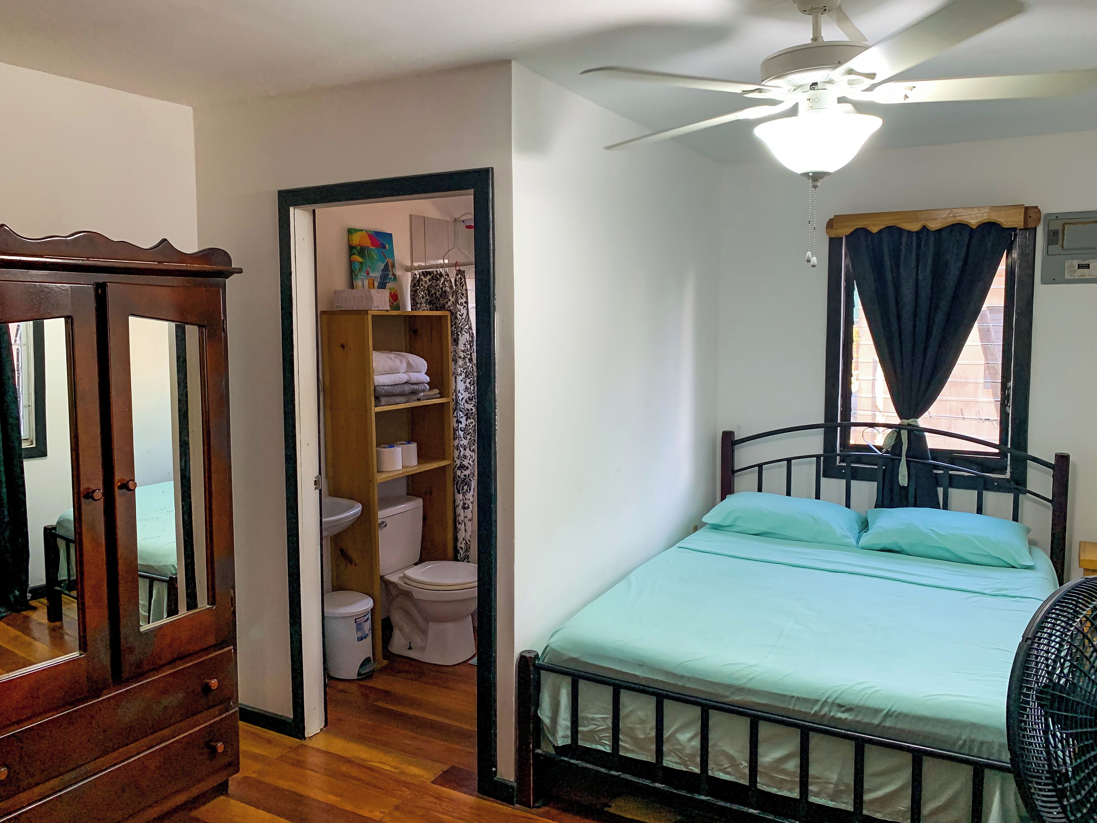 A bedroom with a bed, a ceiling fan, a window with curtains, a wooden wardrobe, and a bathroom with a toilet and shelves.