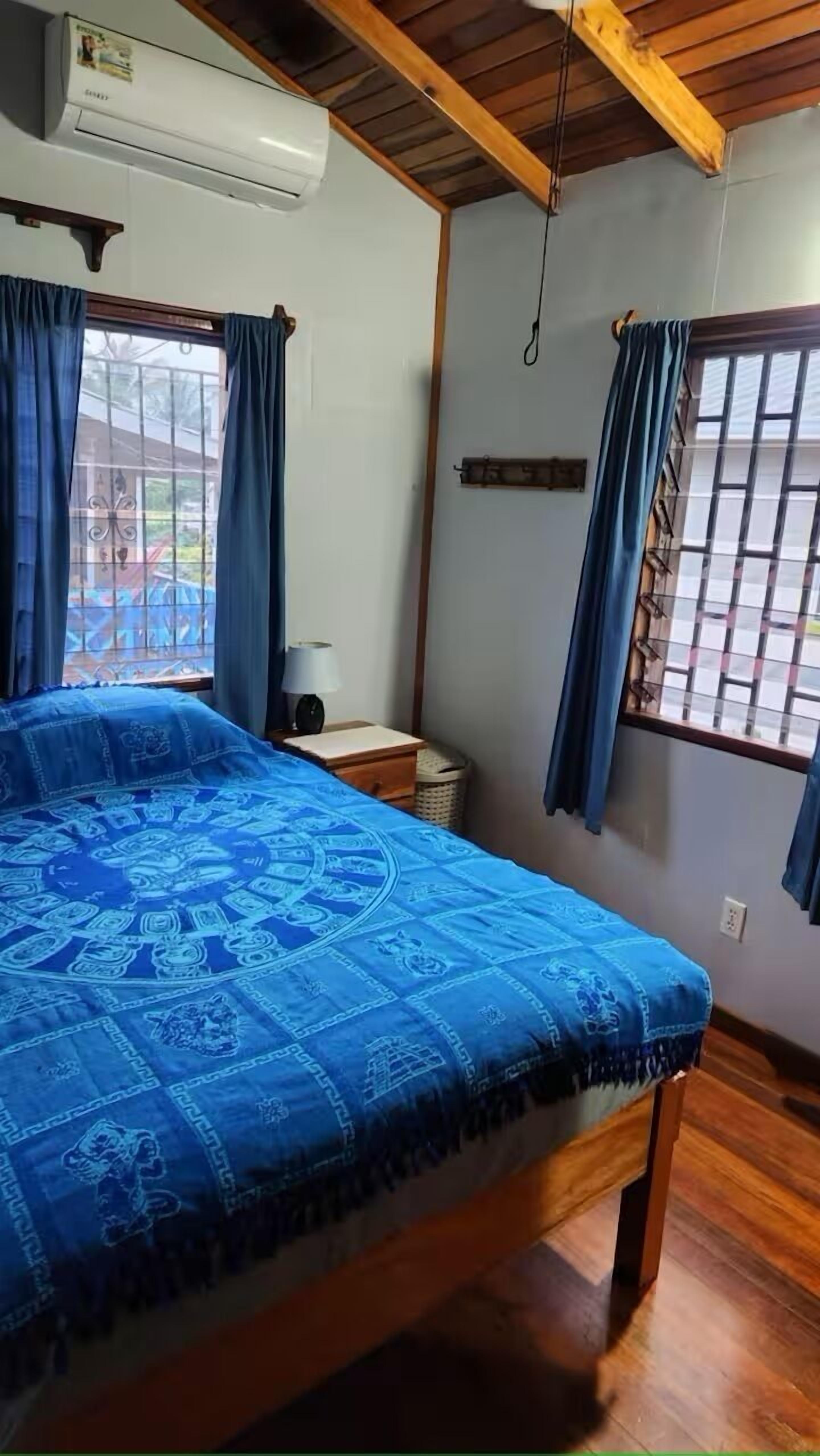 A bedroom with a bed, a nightstand, a lamp, a window with curtains, and an air conditioning unit.