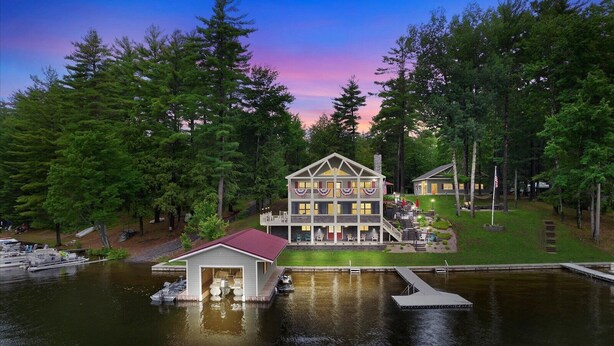 A large, multi-story lodge with a dock and a small boat house, surrounded by dense forest.