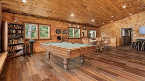 A wooden interior with a pool table, bookshelves, and a dartboard.