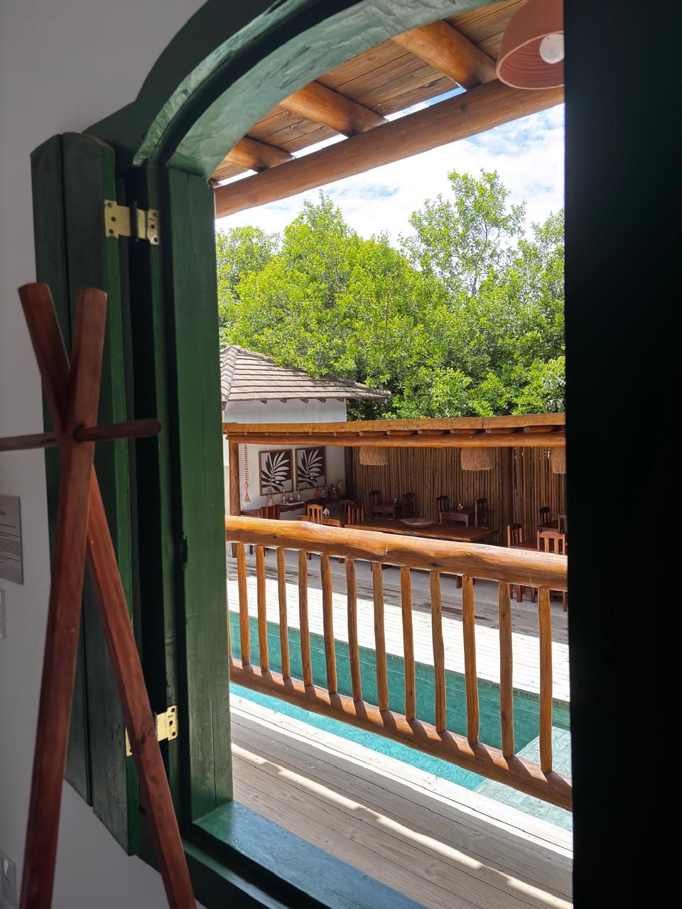 A wooden deck with a railing, a view of a building with a tiled roof, and lush green trees in the background.