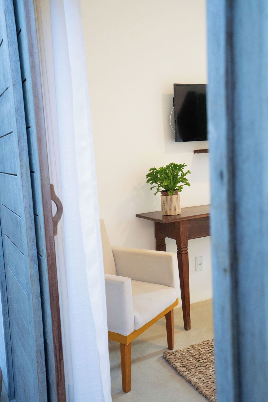 A room with a wooden chair, a small table with a potted plant, and a television mounted on the wall.