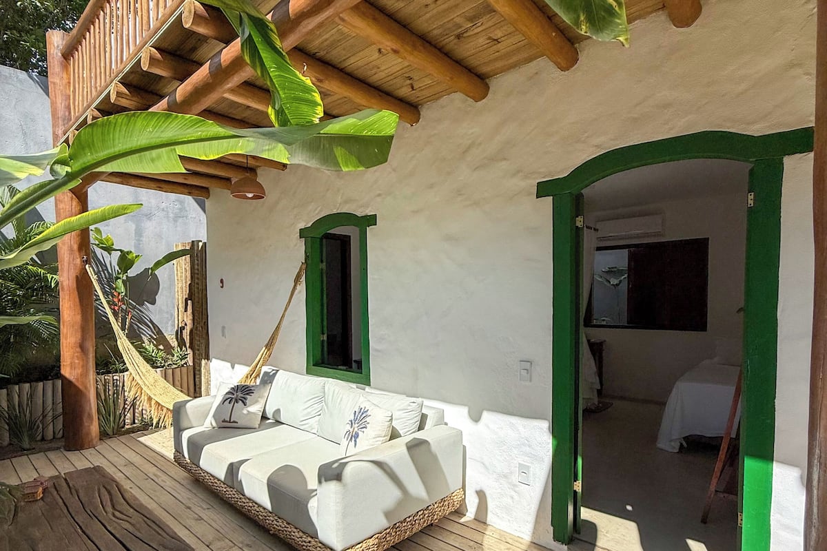 A wooden deck with a white sofa and a hammock, surrounded by lush greenery and a wooden pergola.