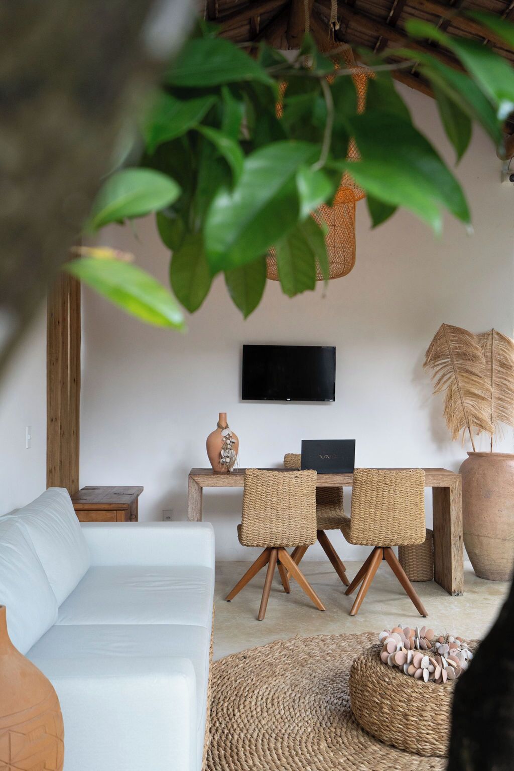 A living room with a white sofa, a dining table with wicker chairs, a TV mounted on the wall, and a large potted plant.