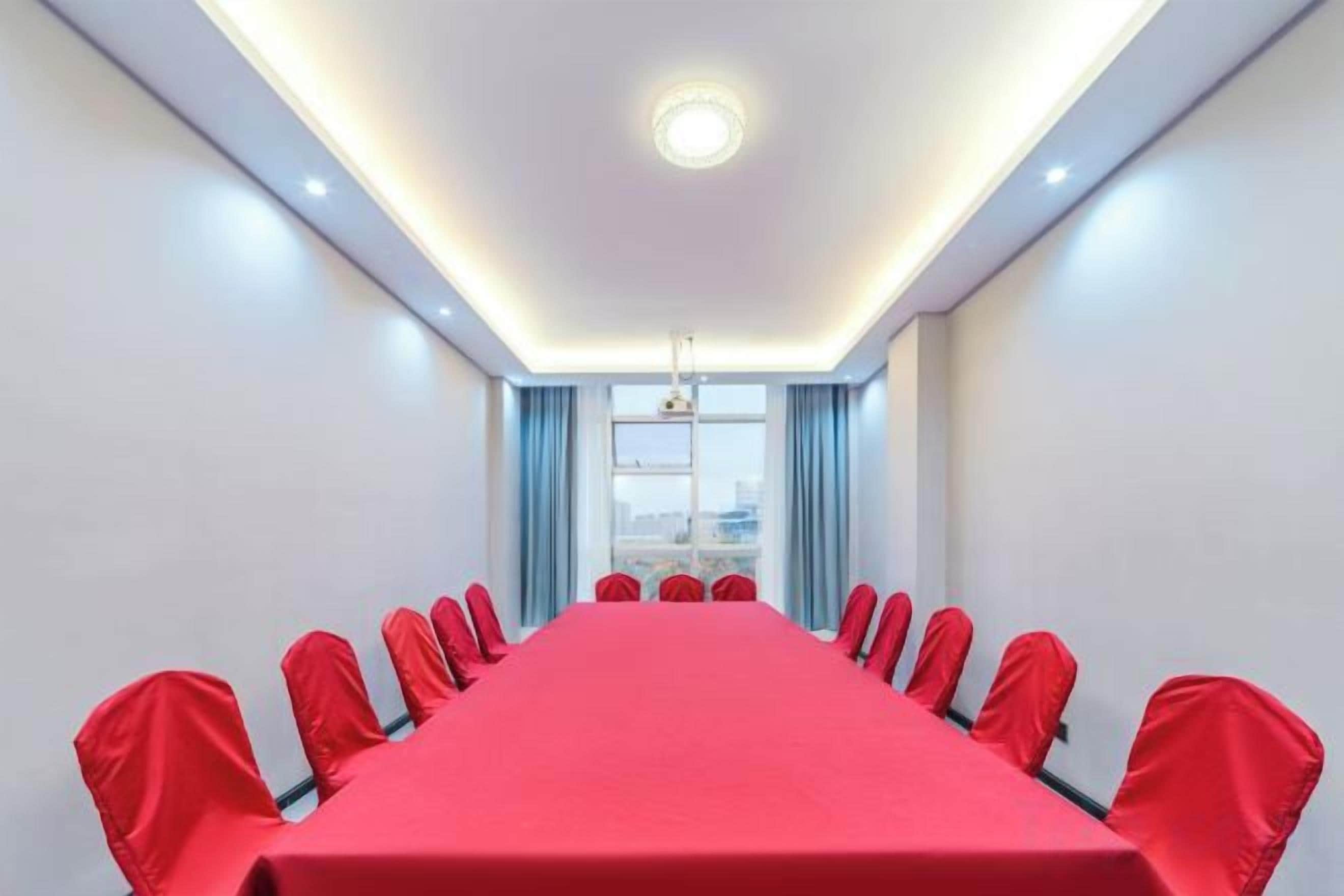 Une salle de conférence avec une longue table et des chaises rouges, un luminaire au plafond et une fenêtre avec des rideaux.