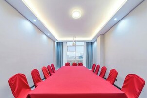Un salón de conferencias con una larga mesa y sillas rojas, una lámpara en el techo y una ventana con cortinas.