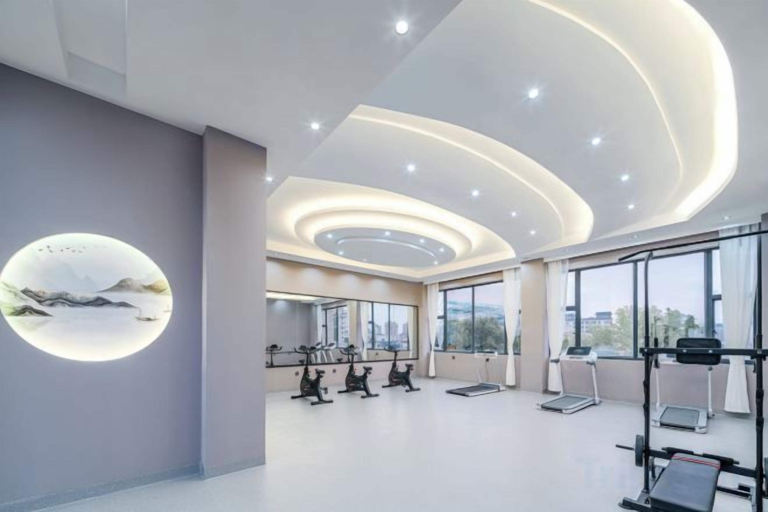 A modern gym with exercise equipment, large windows, and a circular light feature on the wall.