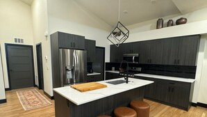 Eine moderne Küche mit dunklen Schränken, einem Edelstahlkühlschrank, einer zentralen Kochinsel mit Sitzgelegenheiten und einer Pendelleuchte.