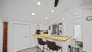 A modern kitchen with a wooden countertop, two bar stools, a stainless steel trash can, and a clock on the wall.