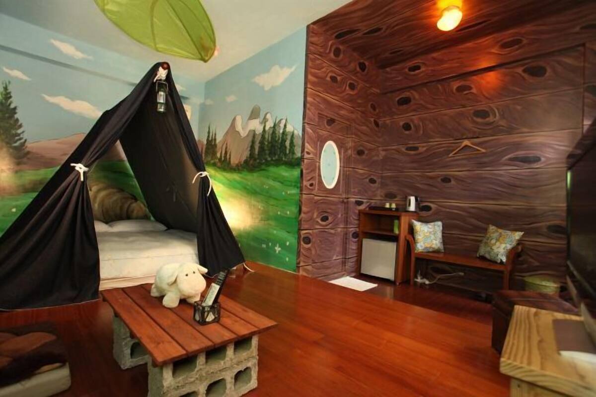Una habitación con pared de madera, una cama con dosel negro, una mesita con un peluche y un televisor.