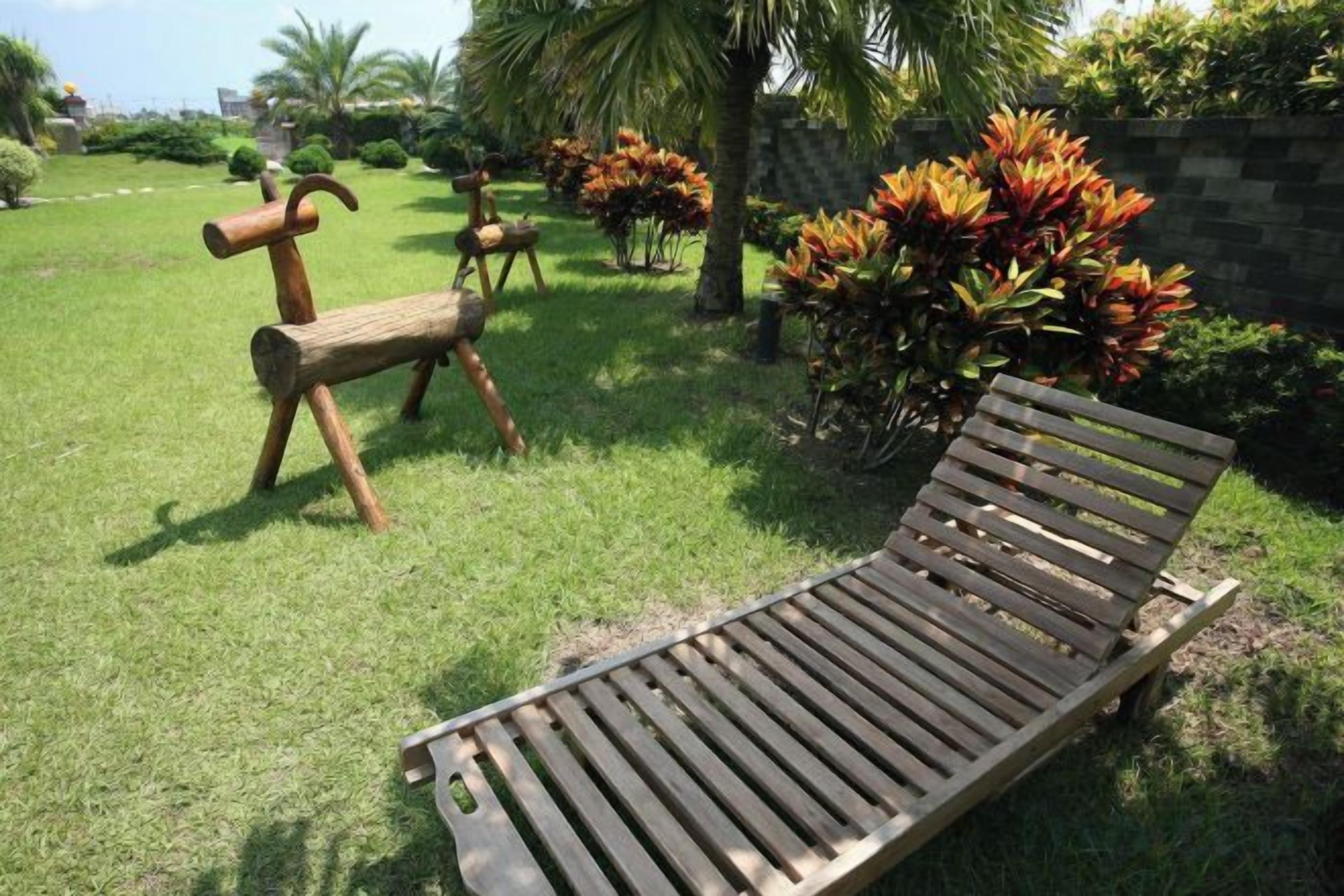 A wooden lounge chair and a rustic wooden bench in a grassy outdoor area with plants and a stone wall.