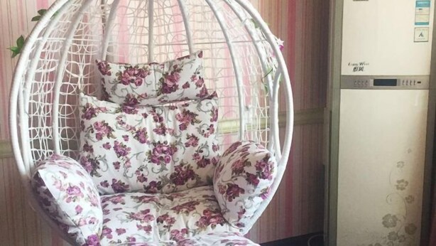 Une chaise suspendue blanche, recouverte d’un tissu à motifs floraux, se trouve dans une pièce dont le papier peint est à rayures.
