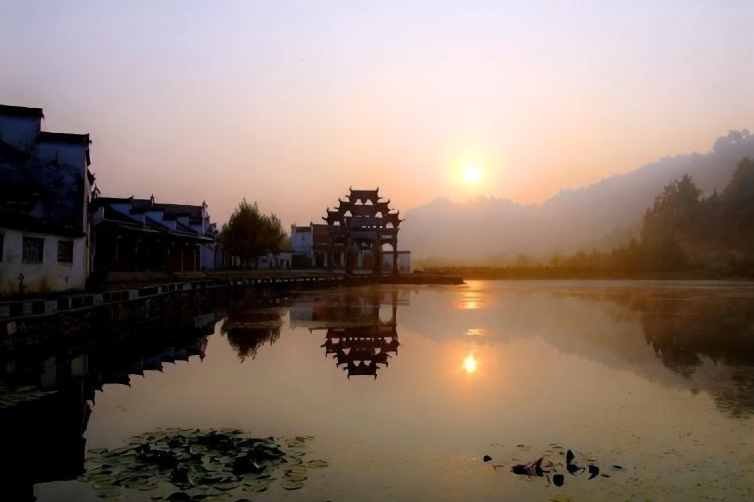 A traditional building with a reflection in a calm body of water, surrounded by a misty landscape.
