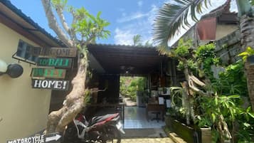 Un espacio al aire libre techado con una motocicleta estacionada, un letrero de bienvenida a Bali y vista hacia un edificio.