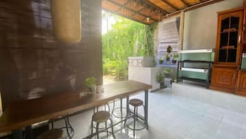 A patio area with a wooden table and stools, a grill, and a view of greenery.