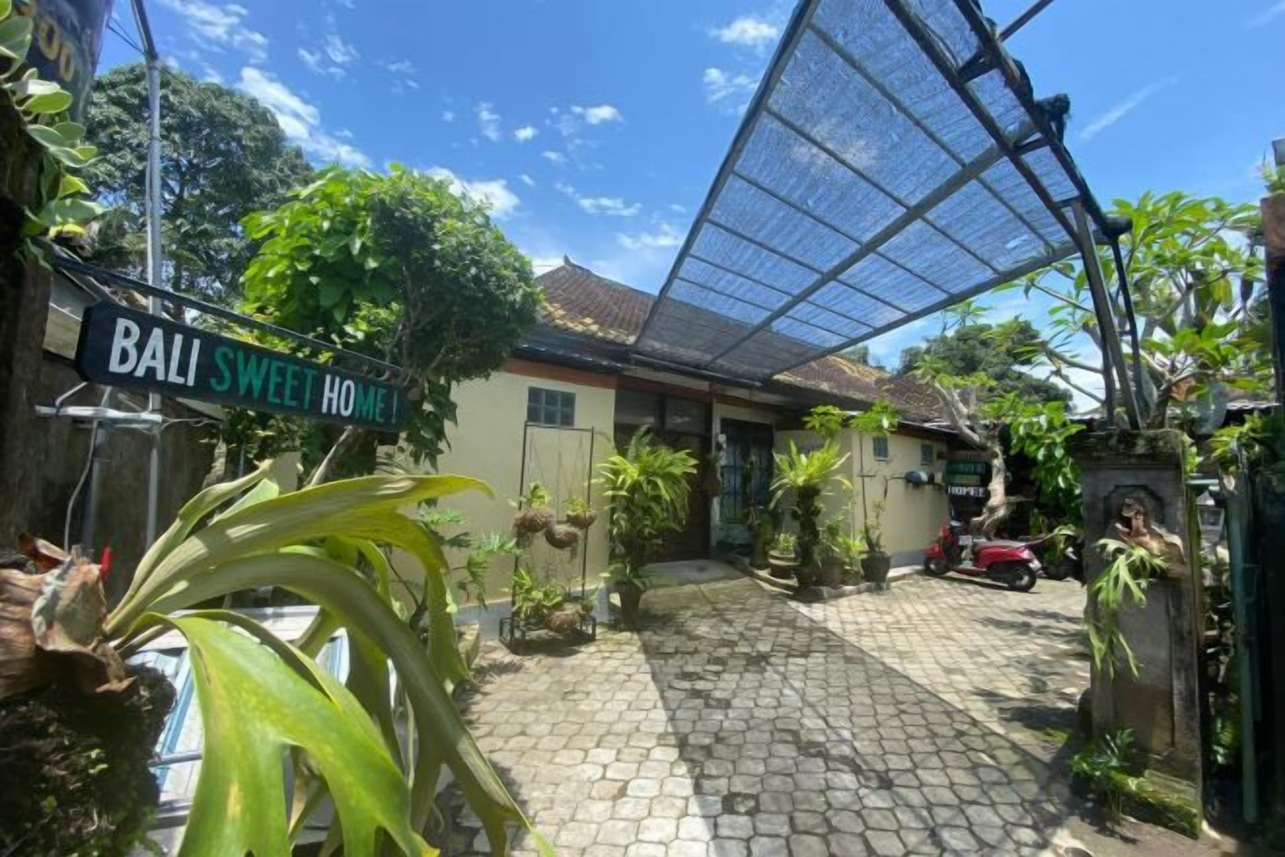 A courtyard with a sign that reads 'BALI SWEET HOME,' surrounded by potted plants and a red vehicle.