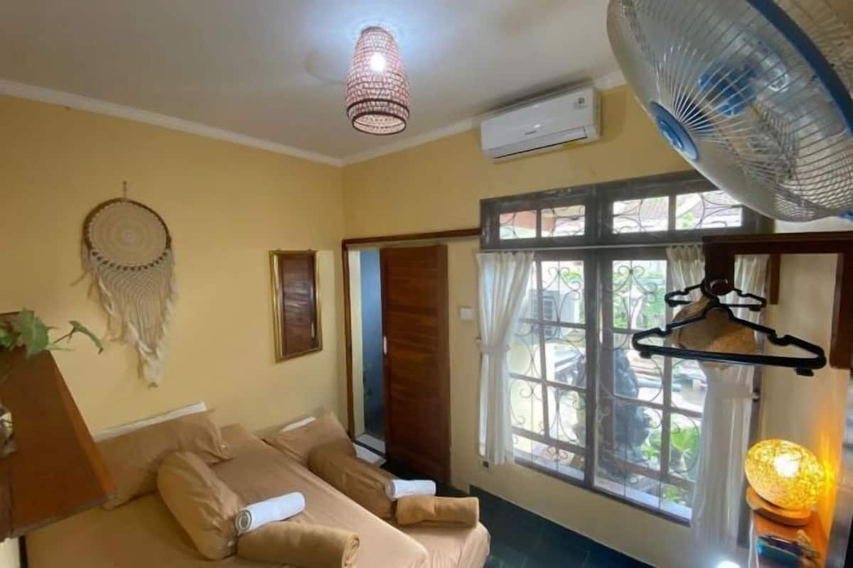 Une pièce avec un lit, un attrape-rêves suspendu, un ventilateur sur pied et une fenêtre avec des rideaux.