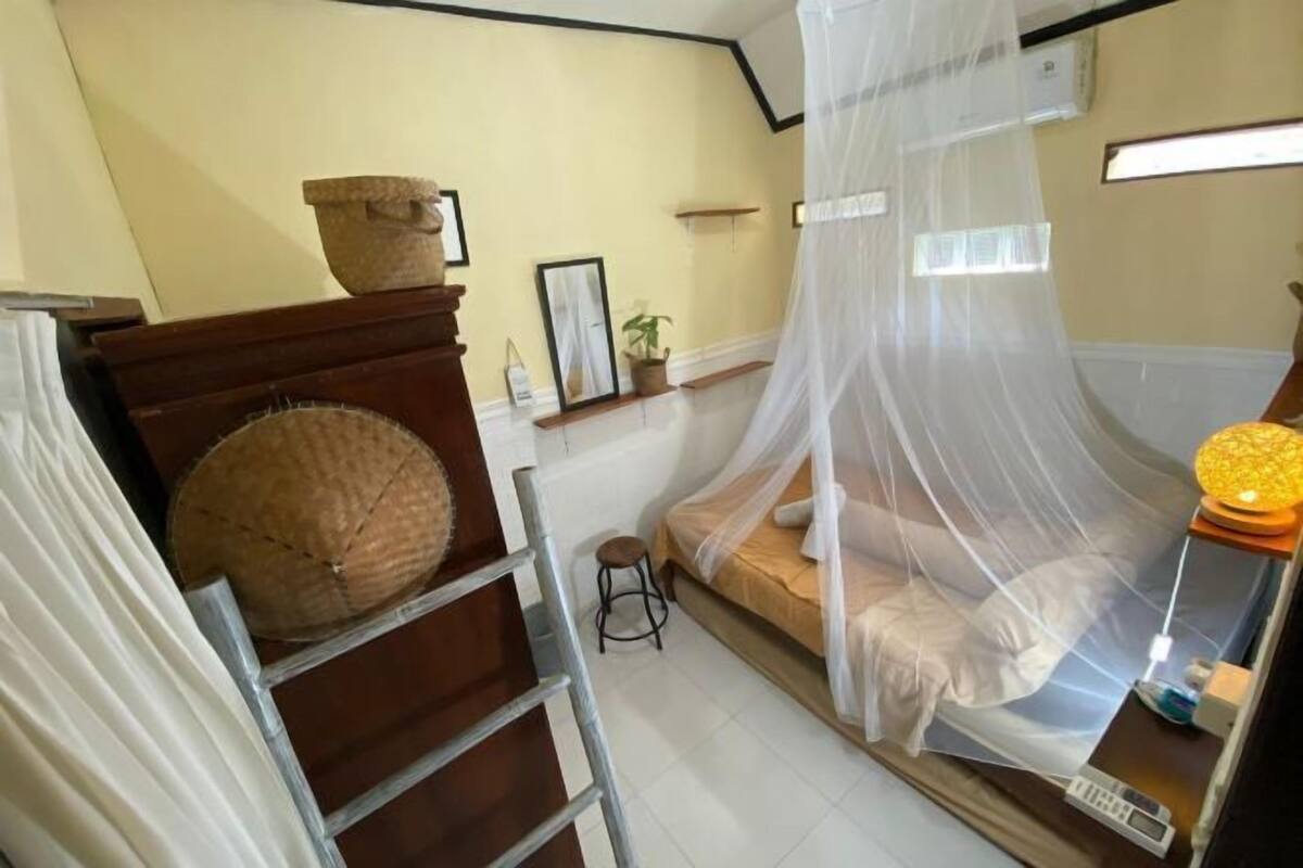 Une chambre à coucher avec un lit recouvert d’une moustiquaire, un petit tabouret, une table de chevet avec une lampe et une fenêtre avec des stores.