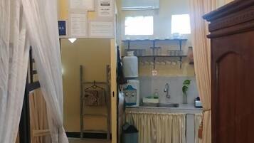 Cocina compacta con dispensador de agua, un lavamanos y una pequeña encimera. Hay una ventana sobre el lavamanos, y se ve un cubo de basura en la esquina.
