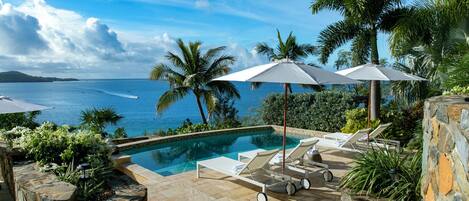A poolside area with lounge chairs and umbrellas, overlooking a body of water and palm trees.