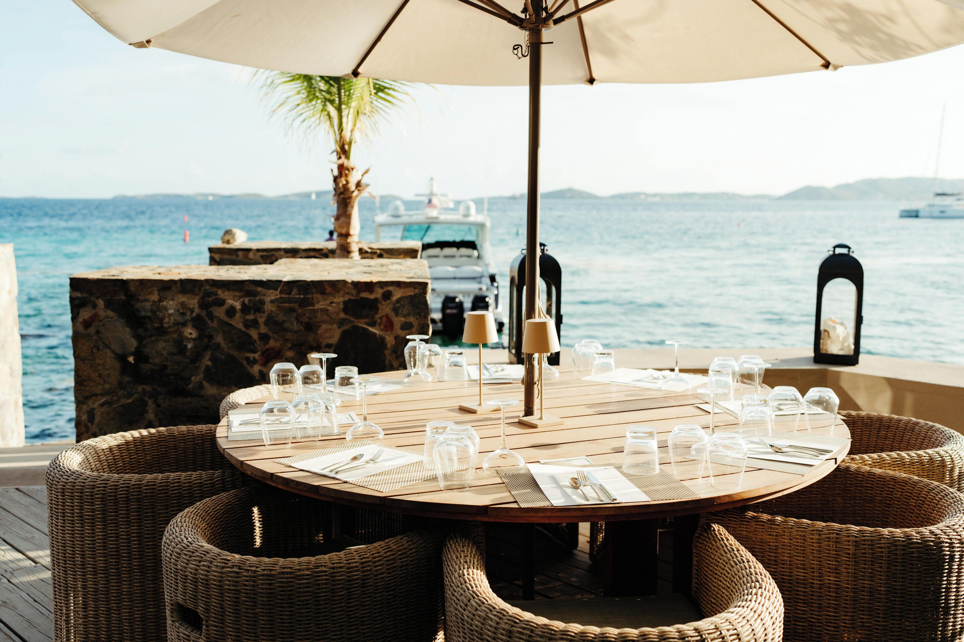 A round wooden table set for dining with wicker chairs, a stone pillar, and a view of the sea with boats in the background.