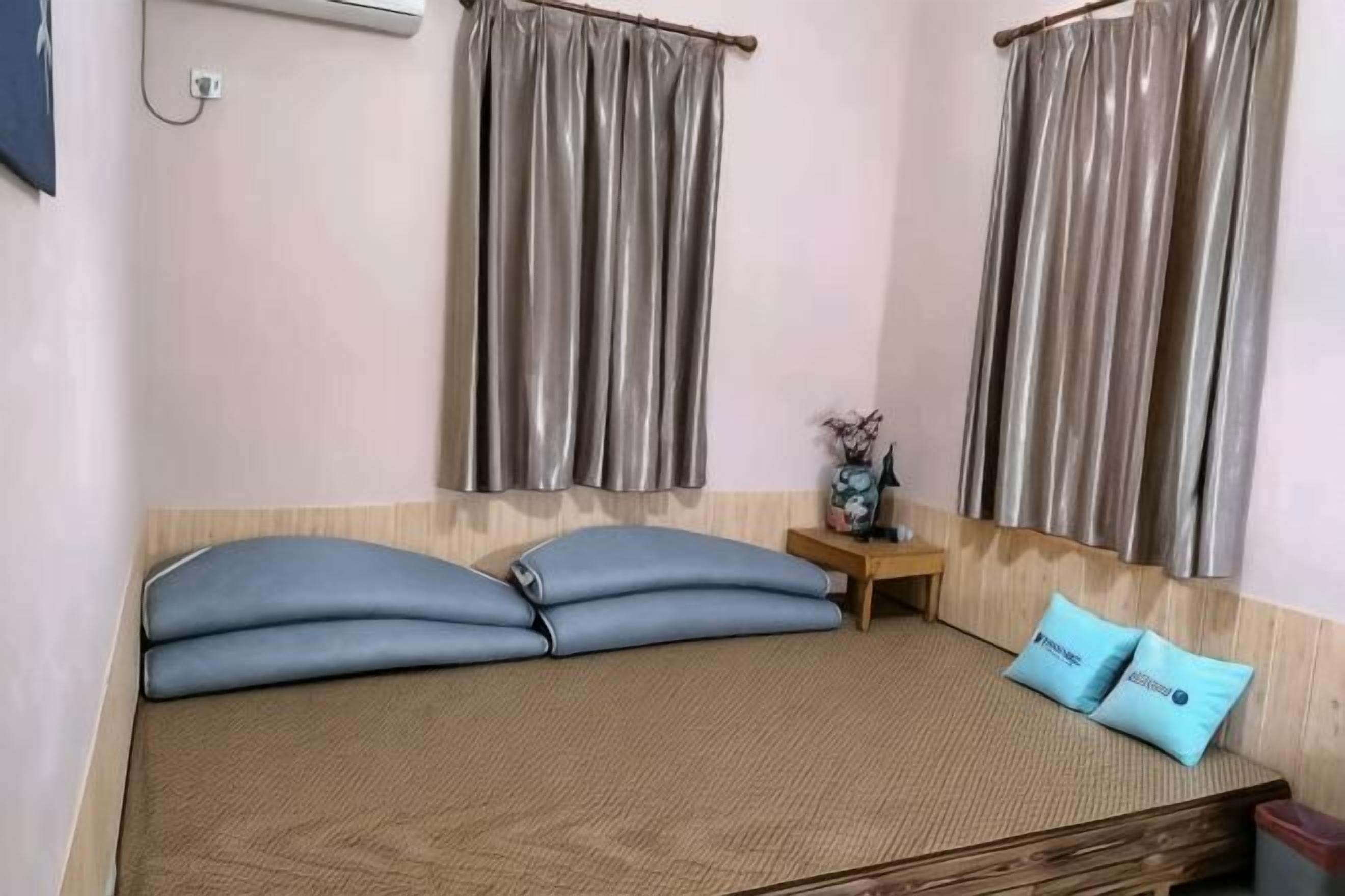 Una habitación con una cama, dos almohadas, una mesita de noche, un florero y cortinas.