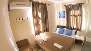 Habitación de hotel con cama, una silla, unidad de aire acondicionado, ventana con cortinas e interruptor de luz en la pared.