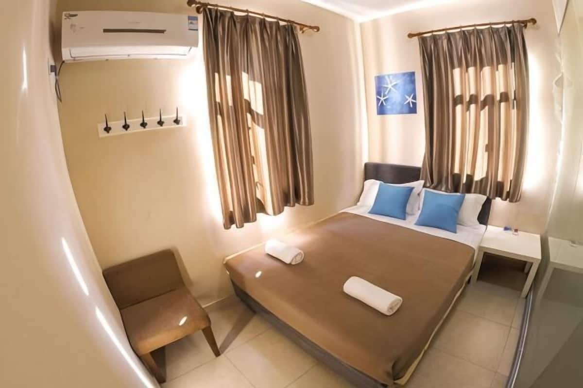 A hotel room with a bed, a chair, an air conditioning unit, a window with curtains, and a wall-mounted light switch.
