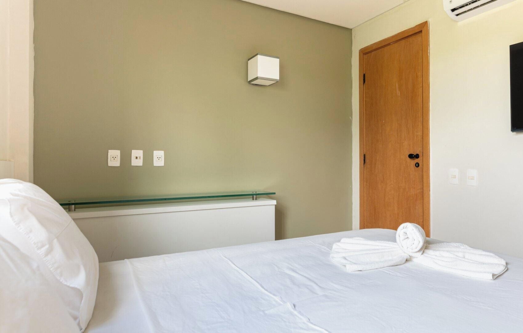A hotel room with a bed, a wooden door, a wall-mounted light, and a glass shelf.