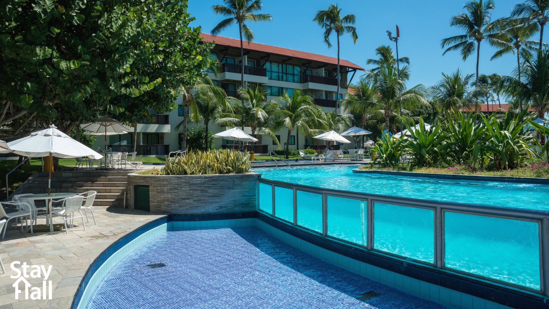 A resort pool area with a clear blue pool, lounge chairs, umbrellas, and palm trees.
