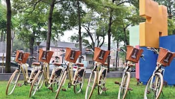 A row of bicycles with baskets, parked on grass with trees and buildings in the background.