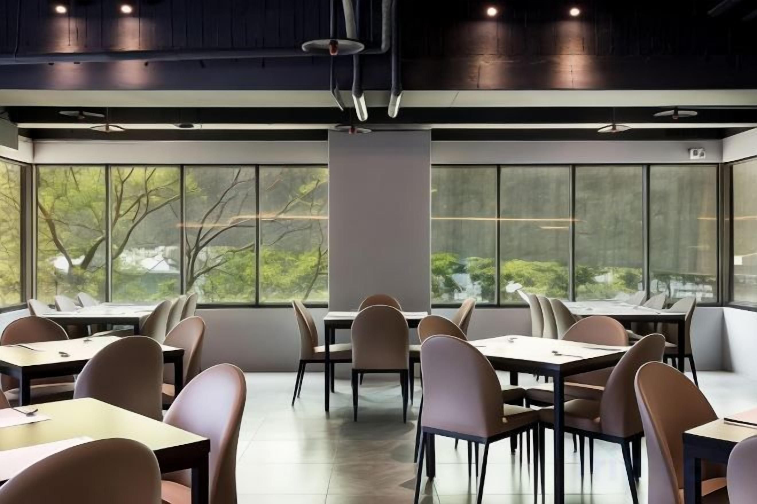 A modern dining area with tables and chairs arranged for groups, large windows providing natural light, and a view of greenery outside.