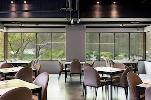 Un comedor moderno con mesas y sillas dispuestas para grupos, ventanas amplias que ofrecen luz natural y vistas a zonas verdes al exterior.