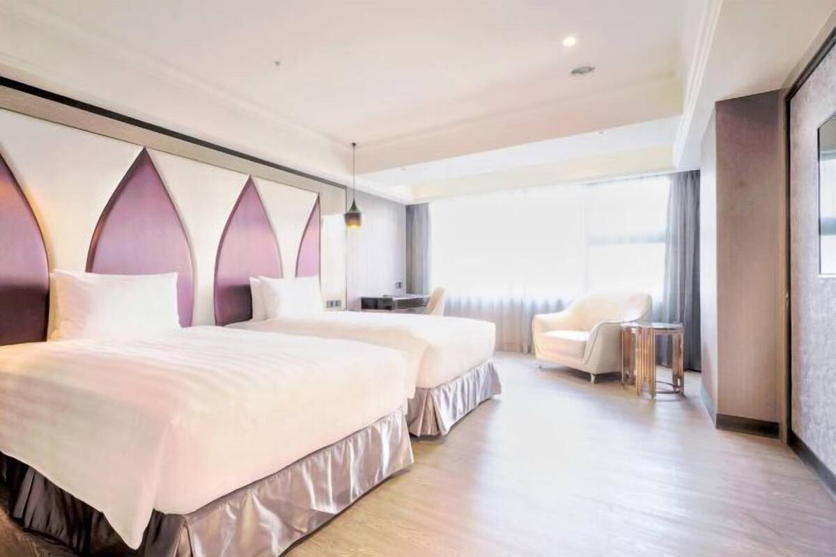 A hotel room with two beds, a desk, a chair, and a large window.
