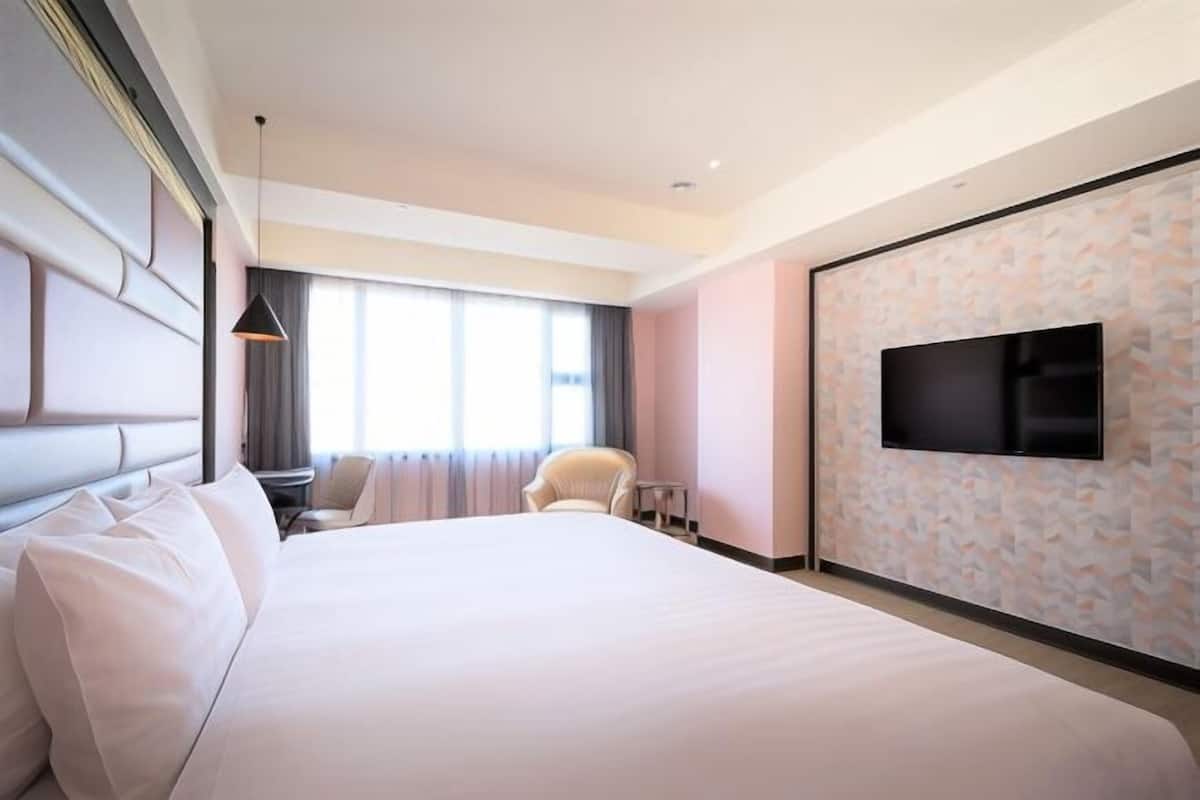 A modern hotel room with a large bed, a flat-screen TV mounted on the wall, a seating area with a chair and a small table, and a large window providing natural light.