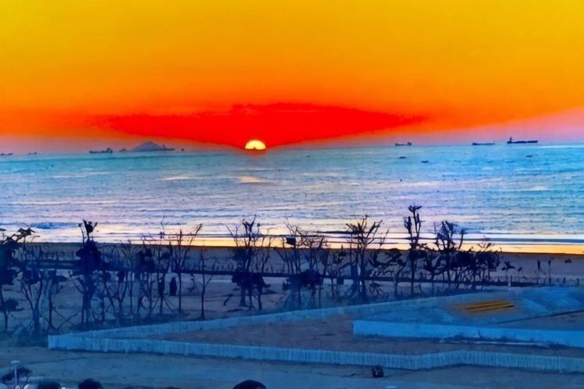 Ein Sonnenuntergang über einem Gewässer, mit Schiffen in der Ferne und einer kargen Landschaft im Vordergrund.