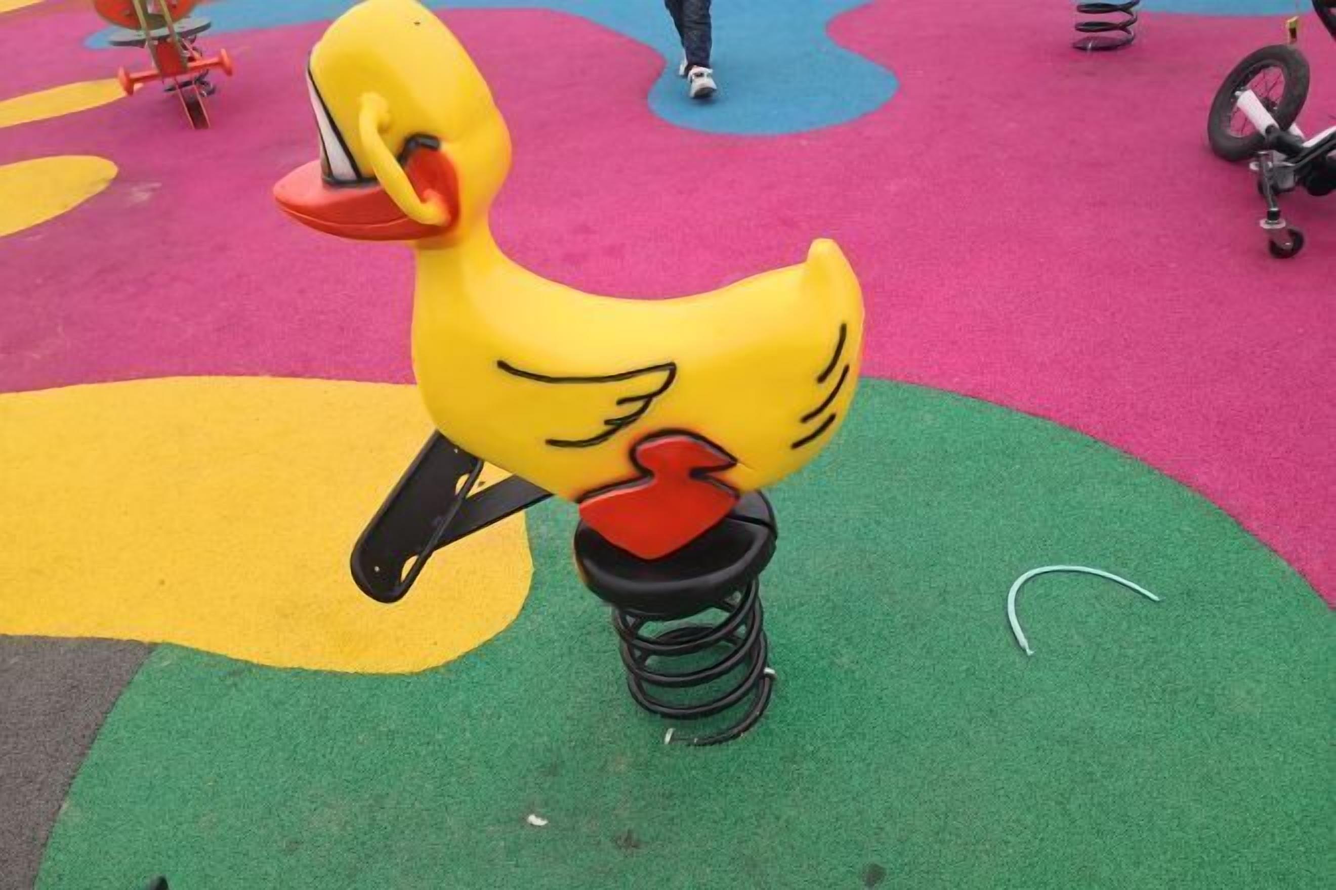 Un columpio con forma de pato amarillo, pico rojo y patas negras, sobre un tapete de juego multicolores.
