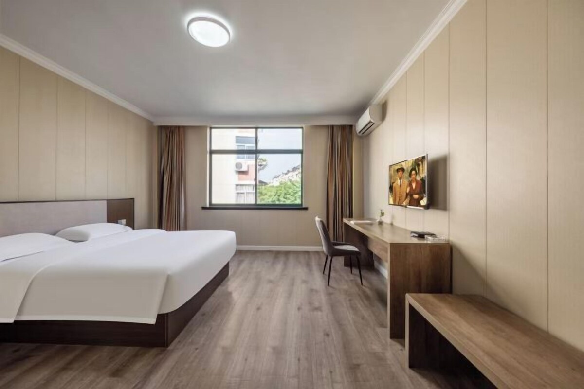 Habitación de hotel con cama, escritorio, silla y un amplio ventanal con vistas a edificios.